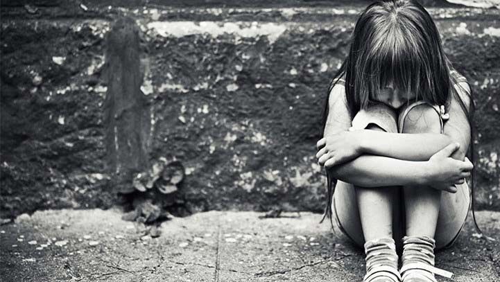 Child huddled on ground with head down, arms wrapped around legs, near a textured wall.
