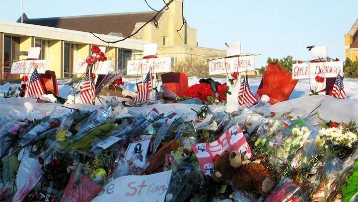 Memorial of flowers, flags, and signs in snow, in front of a building.