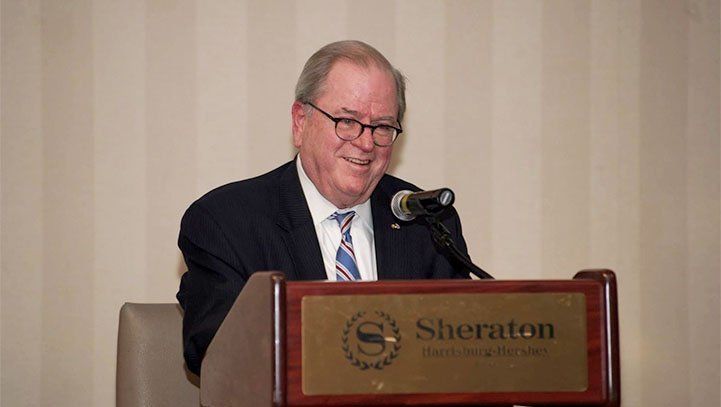 Man speaking at a Sheraton podium, wearing a suit and glasses, smiling.