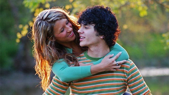 Woman with blonde hair embraces a person with dark curly hair from behind. They are outside with a blurred background.