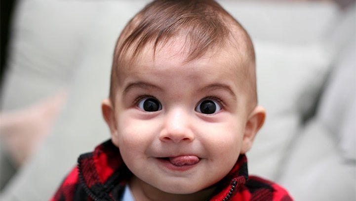 Baby with big eyes and tongue sticking out, wearing a red plaid shirt.