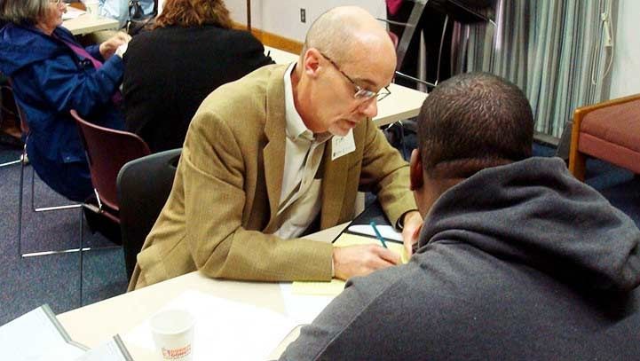 A man in a tan blazer leans over a table, speaking to another person who is taking notes.