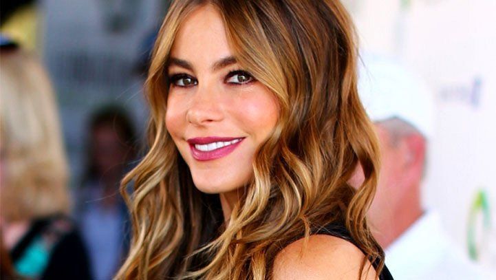 Woman with wavy, highlighted hair smiles, wearing dark lip color; outdoor event background.