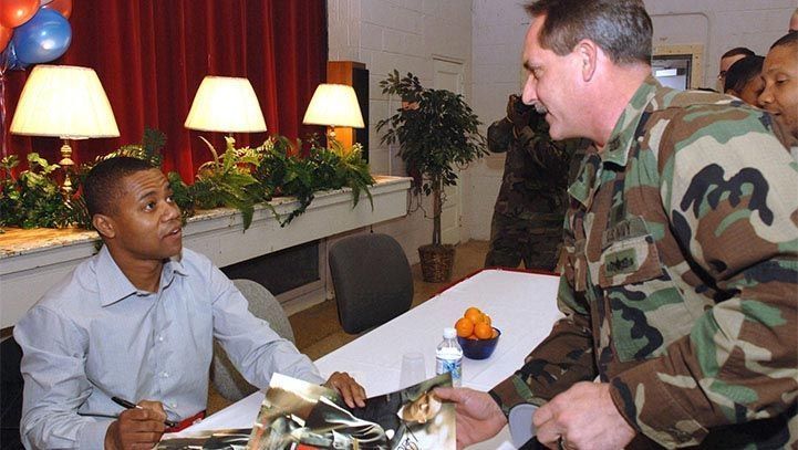 Man signing autographs at a table; soldier in camouflage leans in to converse. Room setting.