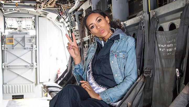 Woman seated in an aircraft, making a peace sign and a kissy face. Wearing a denim jacket and hoodie.