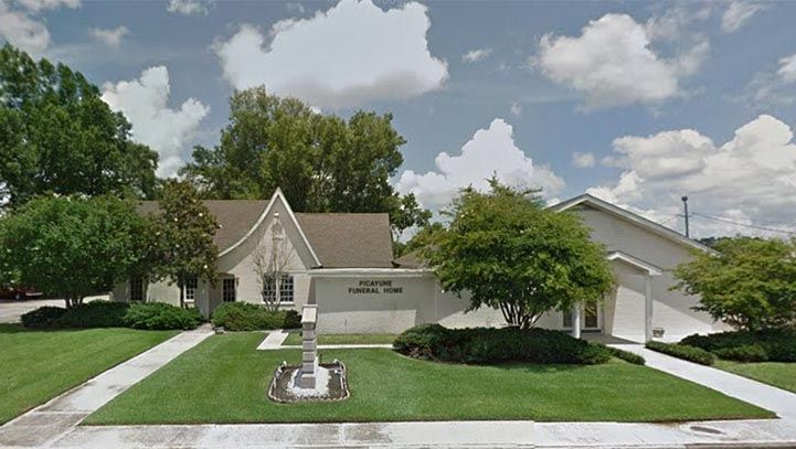 Front exterior of a light-colored funeral home with manicured lawn, cloudy sky.