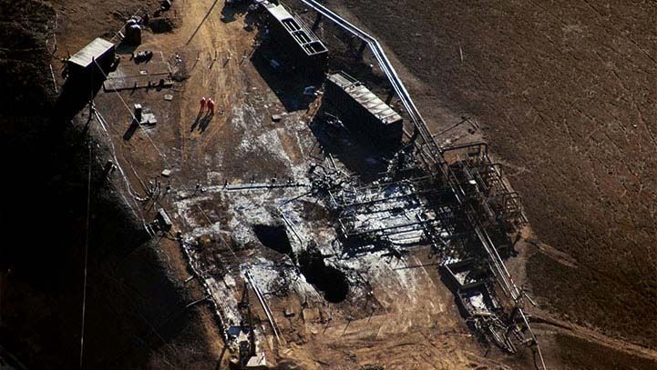 Aerial view of a burned-out building with two people in orange suits standing near the debris, dark setting.