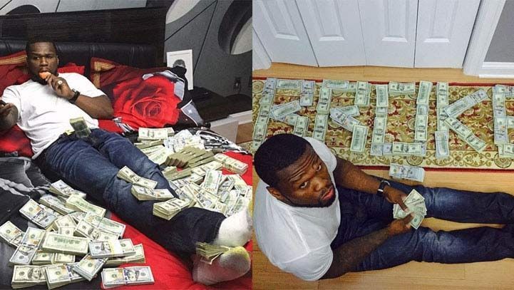 Man surrounded by stacks of money, holding some in his hand on a bed and the floor.