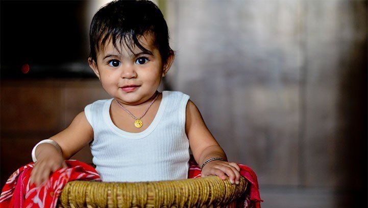 Smiling child in a baby walker, wearing a white shirt and gold necklace, indoor setting.