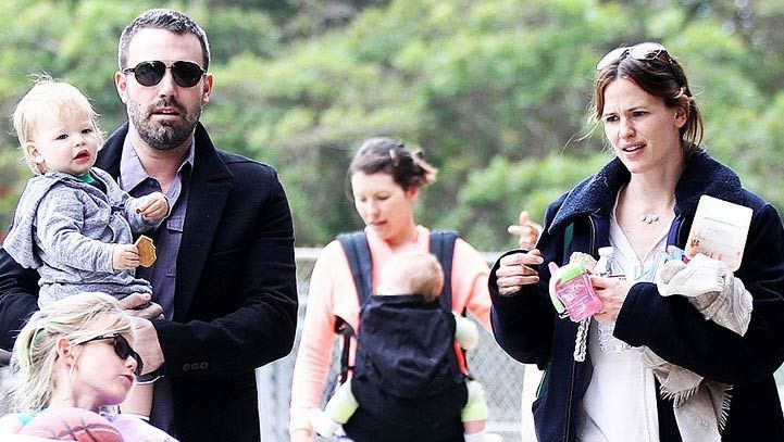 A family walks outdoors: man holding a child, woman holding a baby, another woman carrying a baby.