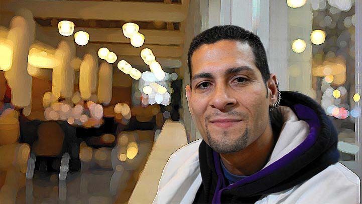 Man in a white jacket smiles, blurred cafe background with lights.