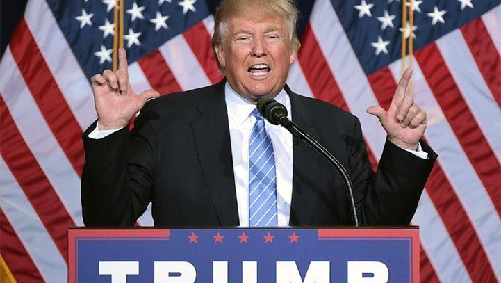 Donald Trump speaking at a podium in front of American flags, gesturing with hands.