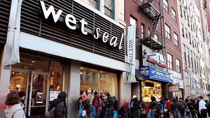 Wet Seal store front, large white logo. People line up in front of a brick building on a city street.