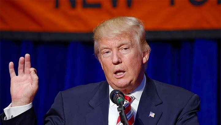 Donald Trump speaking at a podium, gesturing with his hand; blue suit, red tie.