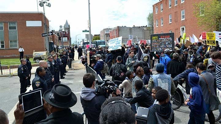Police officers line the street, facing a crowd of protestors holding signs, a news crew present.