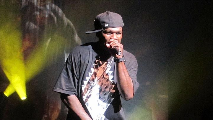 Rapper on stage, wearing a cap and black shirt, singing into a microphone. Yellow stage lights in background.