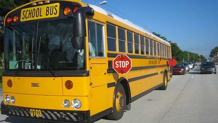 Yellow school bus with stop sign extended on a road, cars stopped behind it on a sunny day.