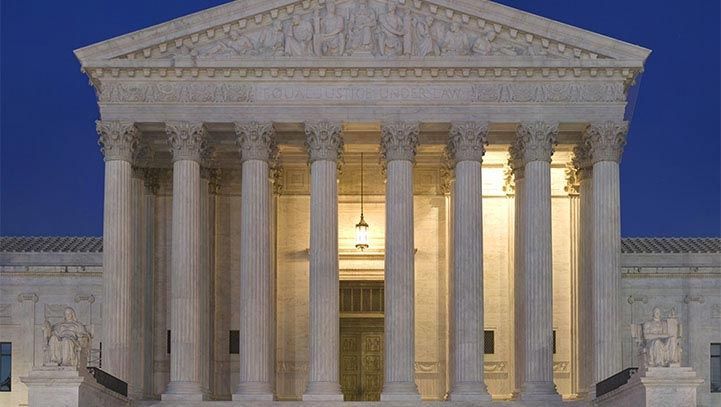 Exterior of the U.S. Supreme Court building at dusk, white columns and facade illuminated against a blue sky.