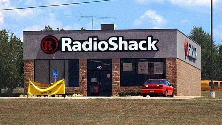 RadioShack store with red sign, red car parked outside, and yellow banner. Building has brick and grey facade.