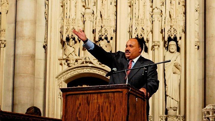 Man speaking from a wooden podium, raising his arm in a church with ornate stone carvings.