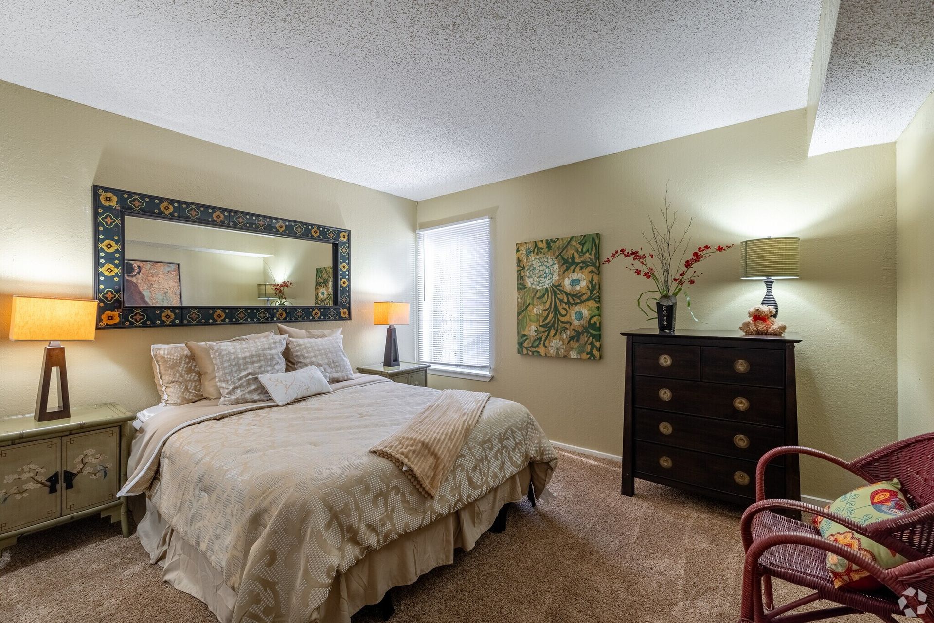 Bedroom with bed, dresser, lamps, window, and wicker chair; beige walls and patterned carpet.