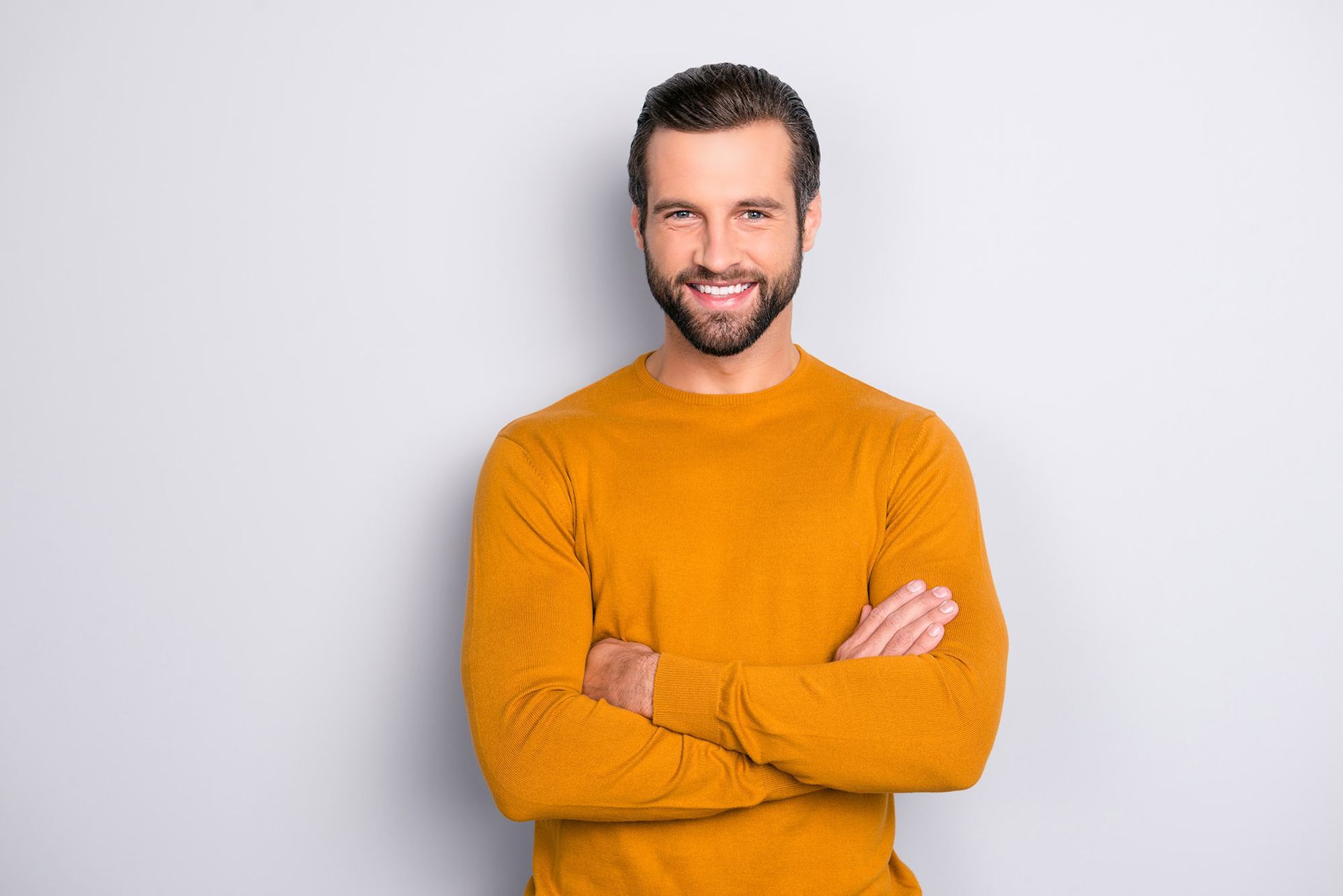 Man with crossed arms smiles, wearing a yellow sweater, against a gray background.