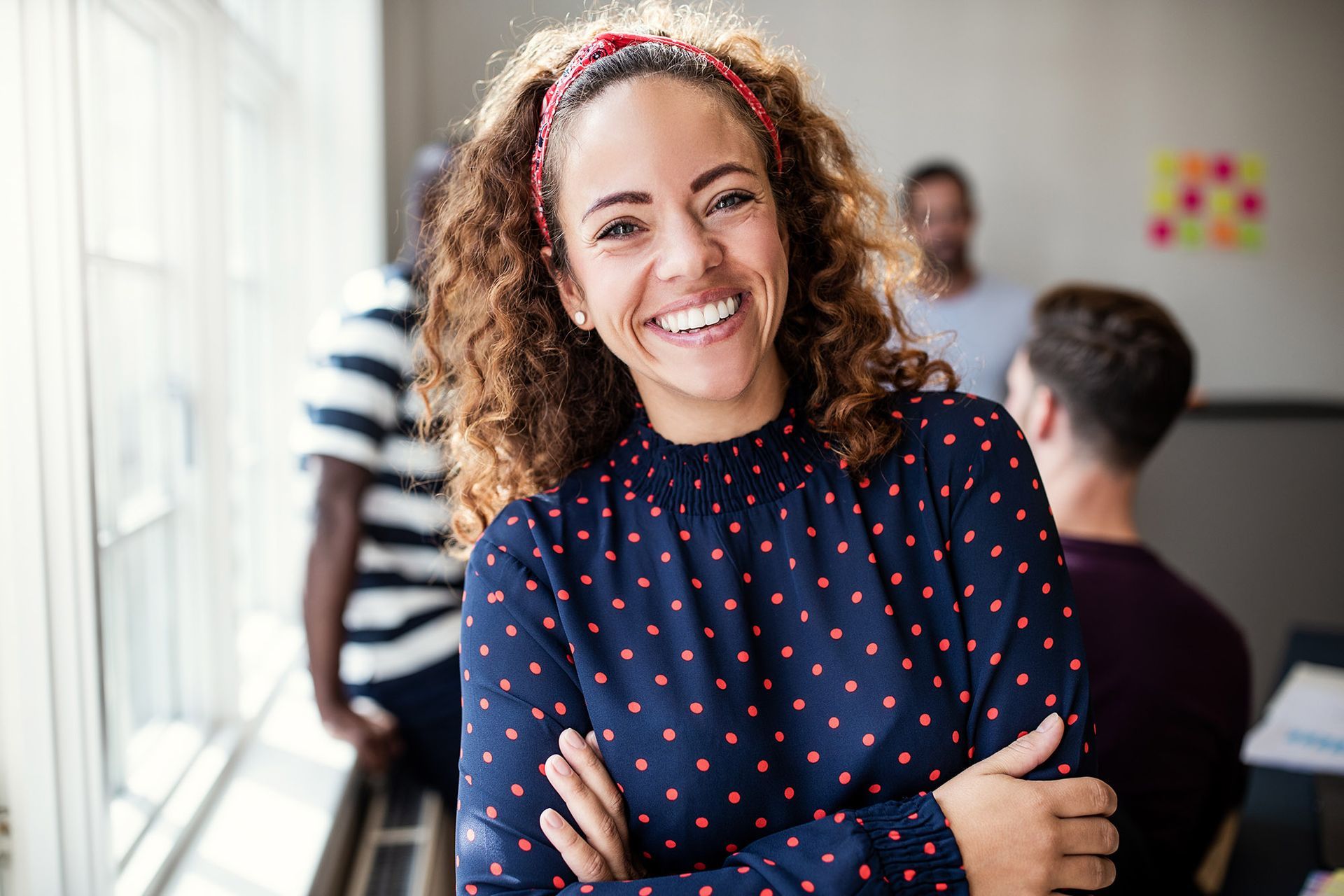 Woman with curly hair smiles, arms crossed, in office setting with coworkers.