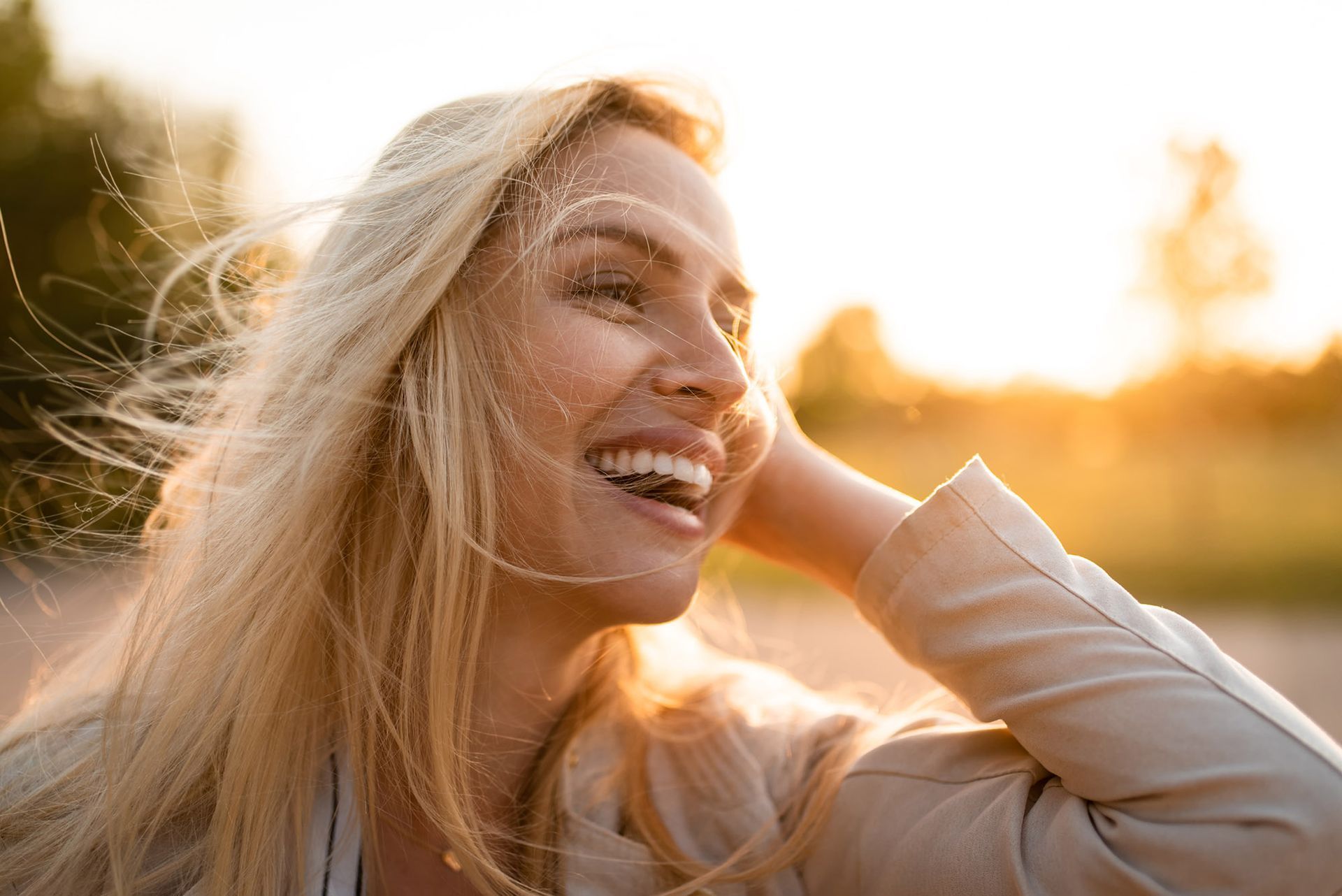 Woman with blonde hair smiling, hand in hair, with sunlit background.