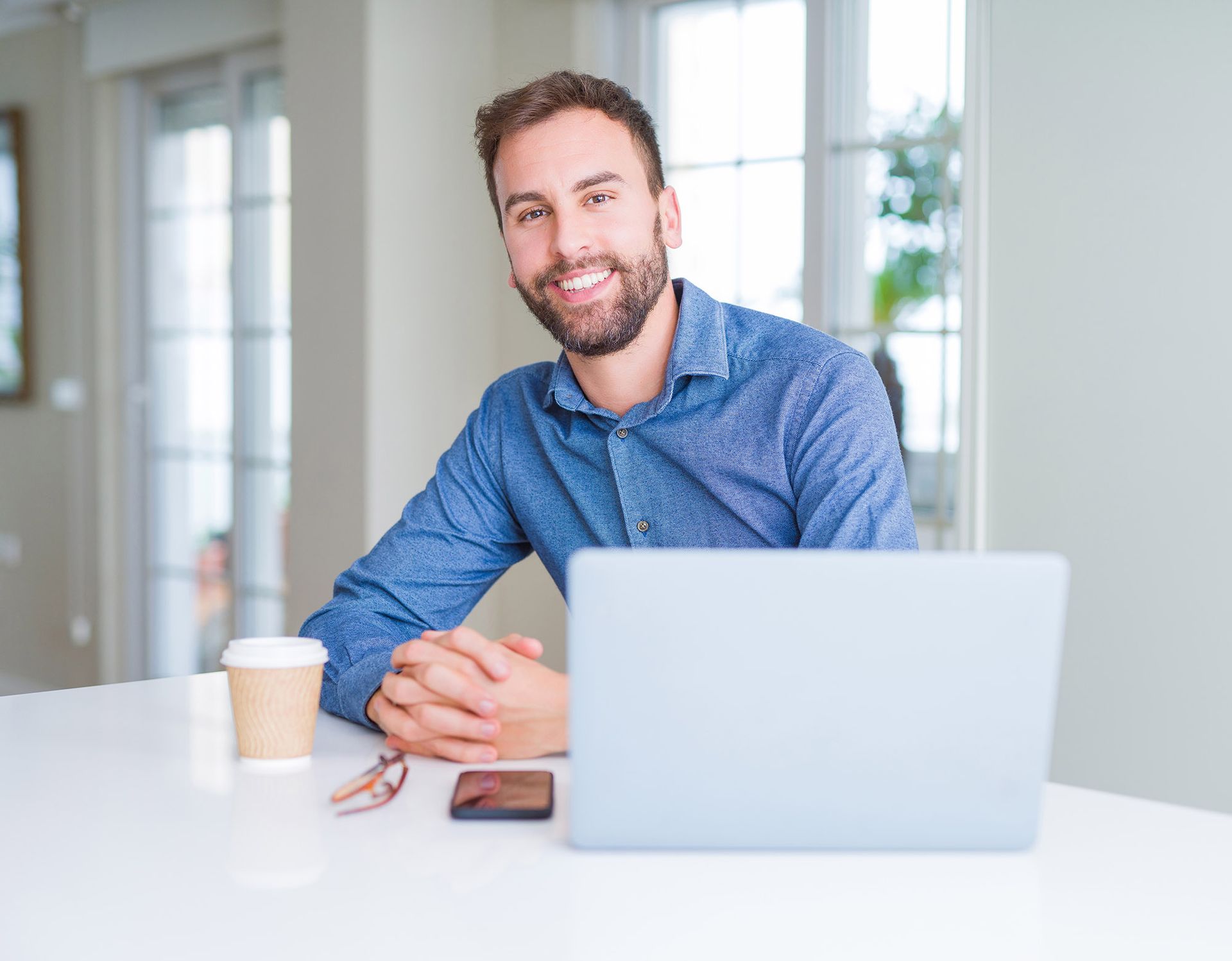 Man smiling at camera while working on a laptop at a table, coffee and phone present.
