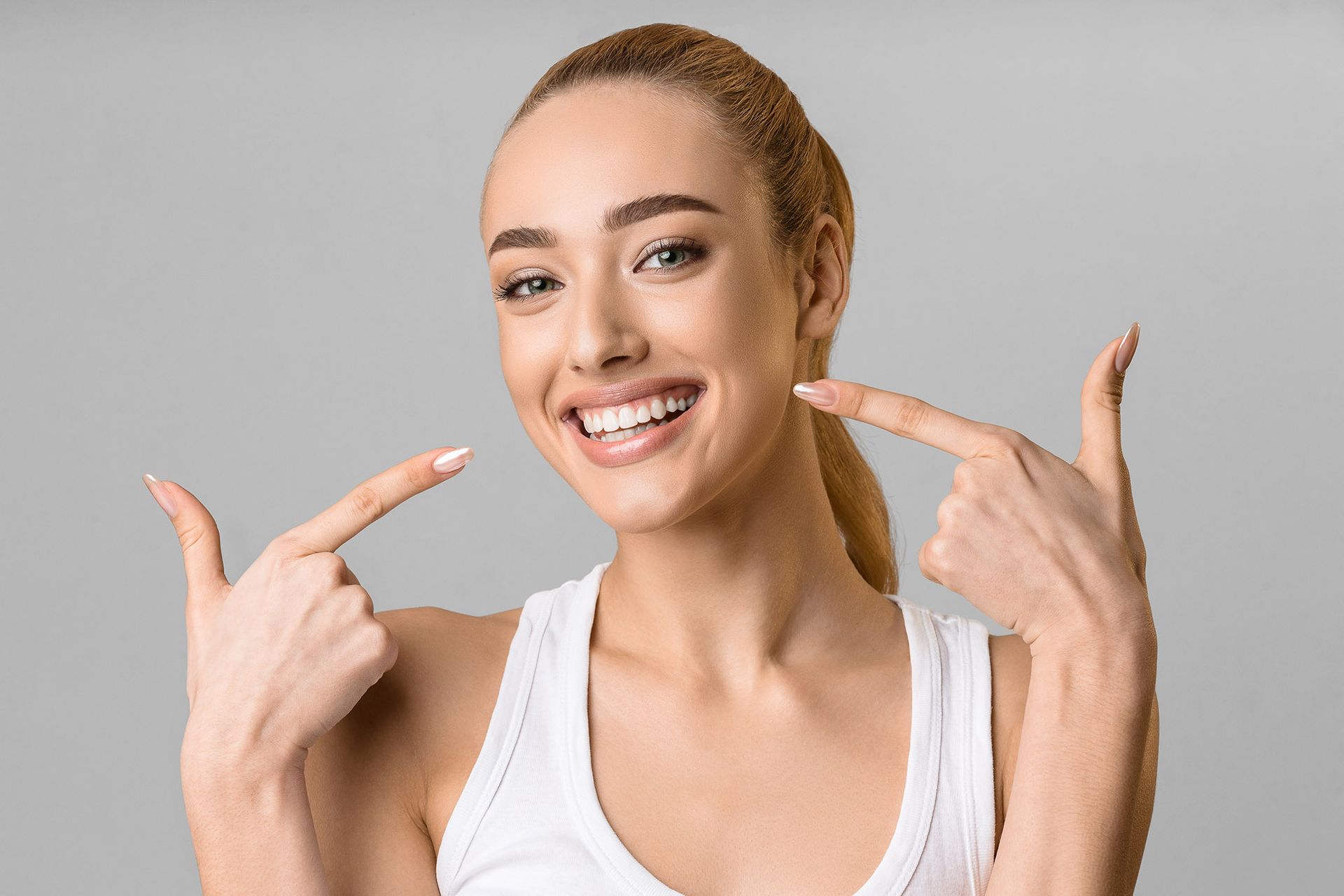 Woman smiling, pointing to her teeth. White tank top, light brown hair pulled back, gray background.