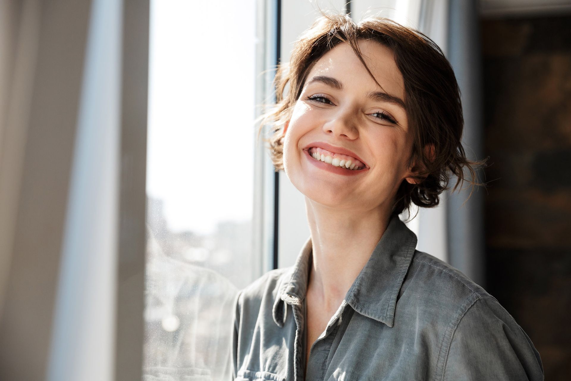 Woman with short brown hair smiling near a window, wearing a button-down shirt.