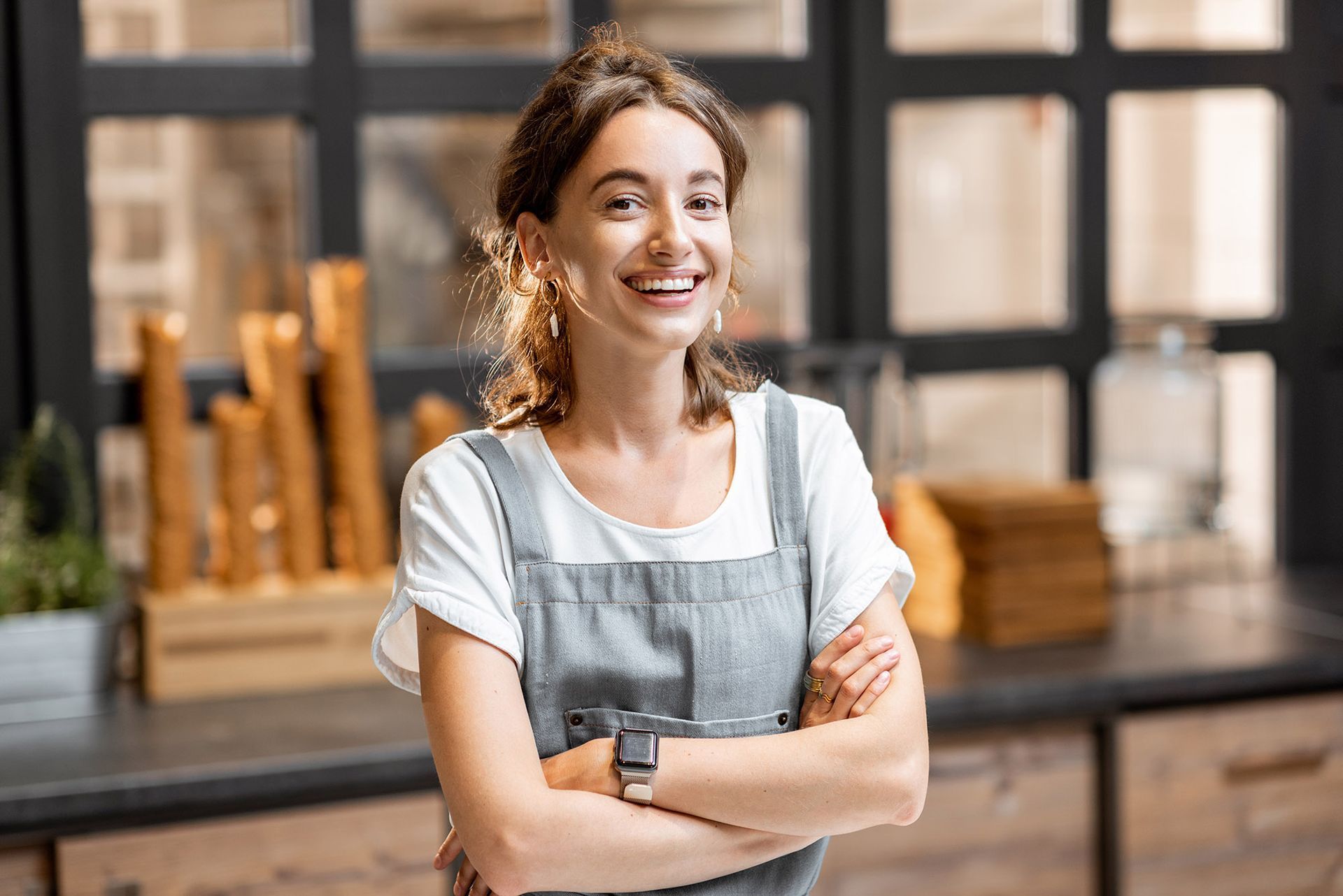 Woman in apron smiles with arms crossed, standing in a cafe.