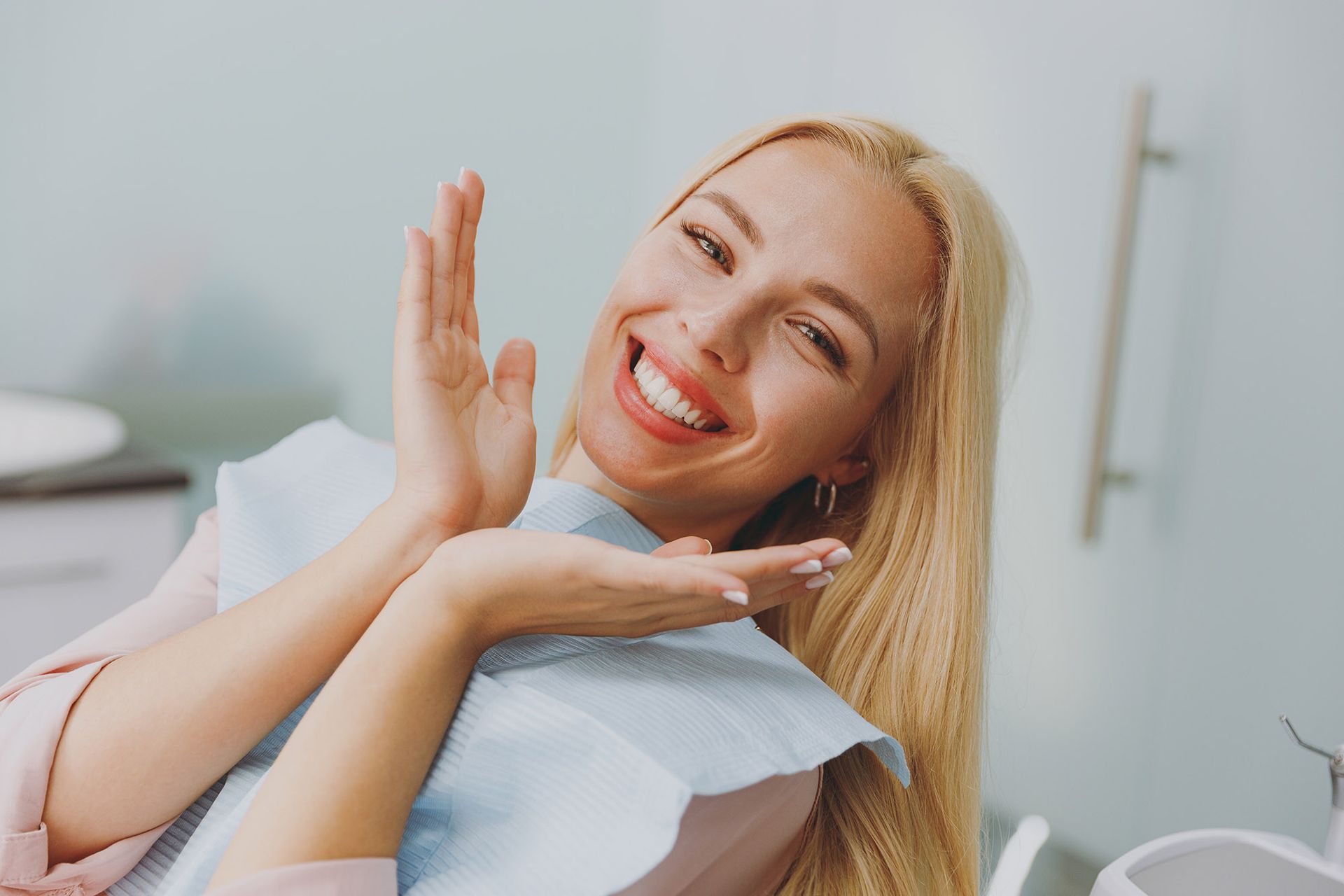 Woman with bright smile in dental chair, gesturing towards her teeth.