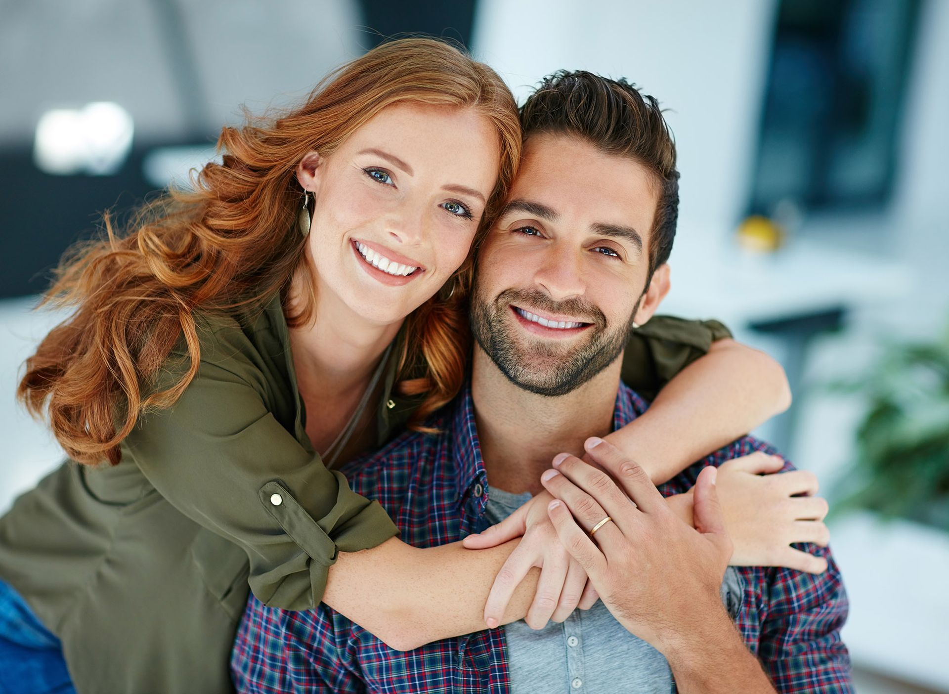Woman with red hair embraces a smiling man with a beard; they are indoors.