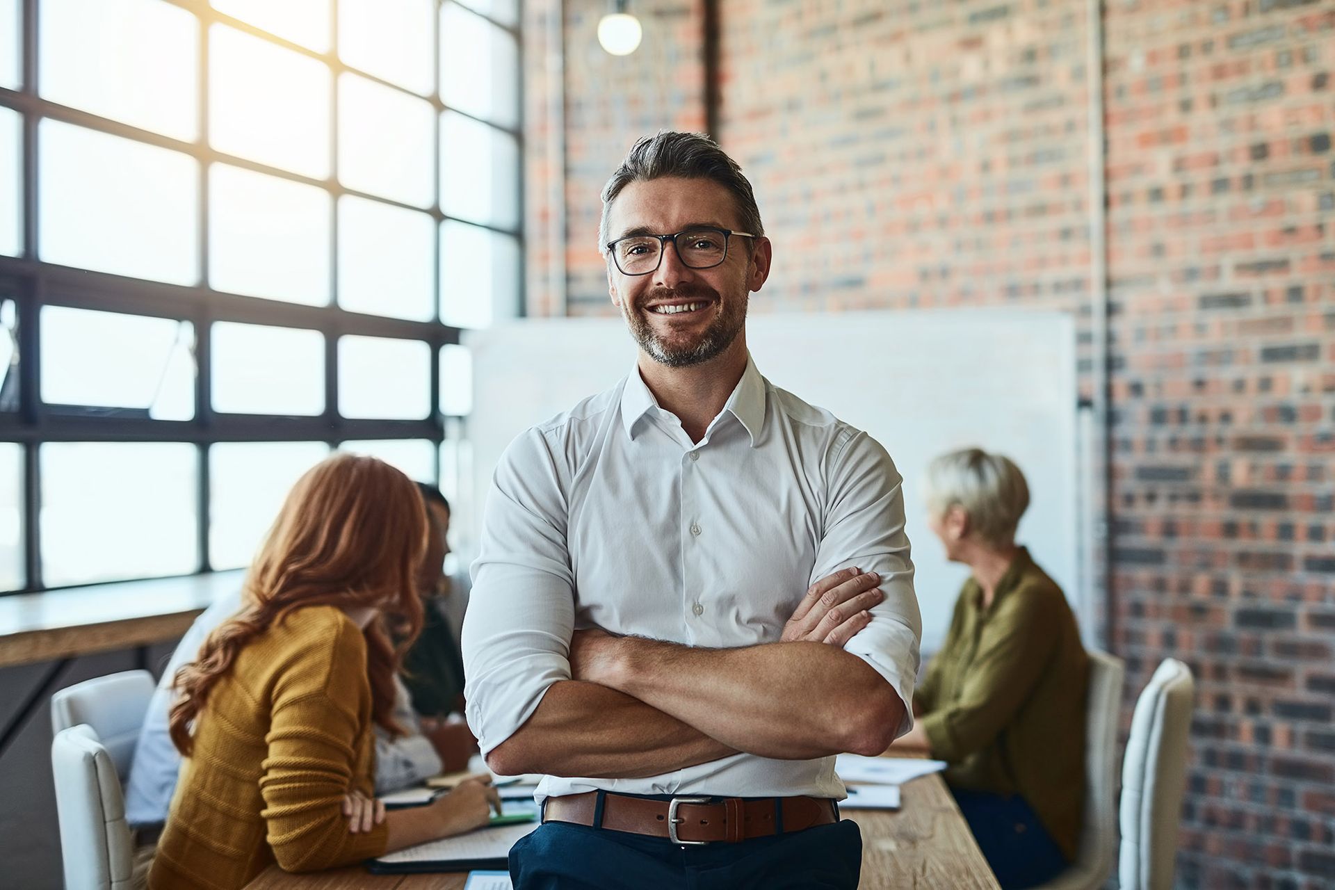 Man with crossed arms smiles in a meeting room, people in the background, brick wall.