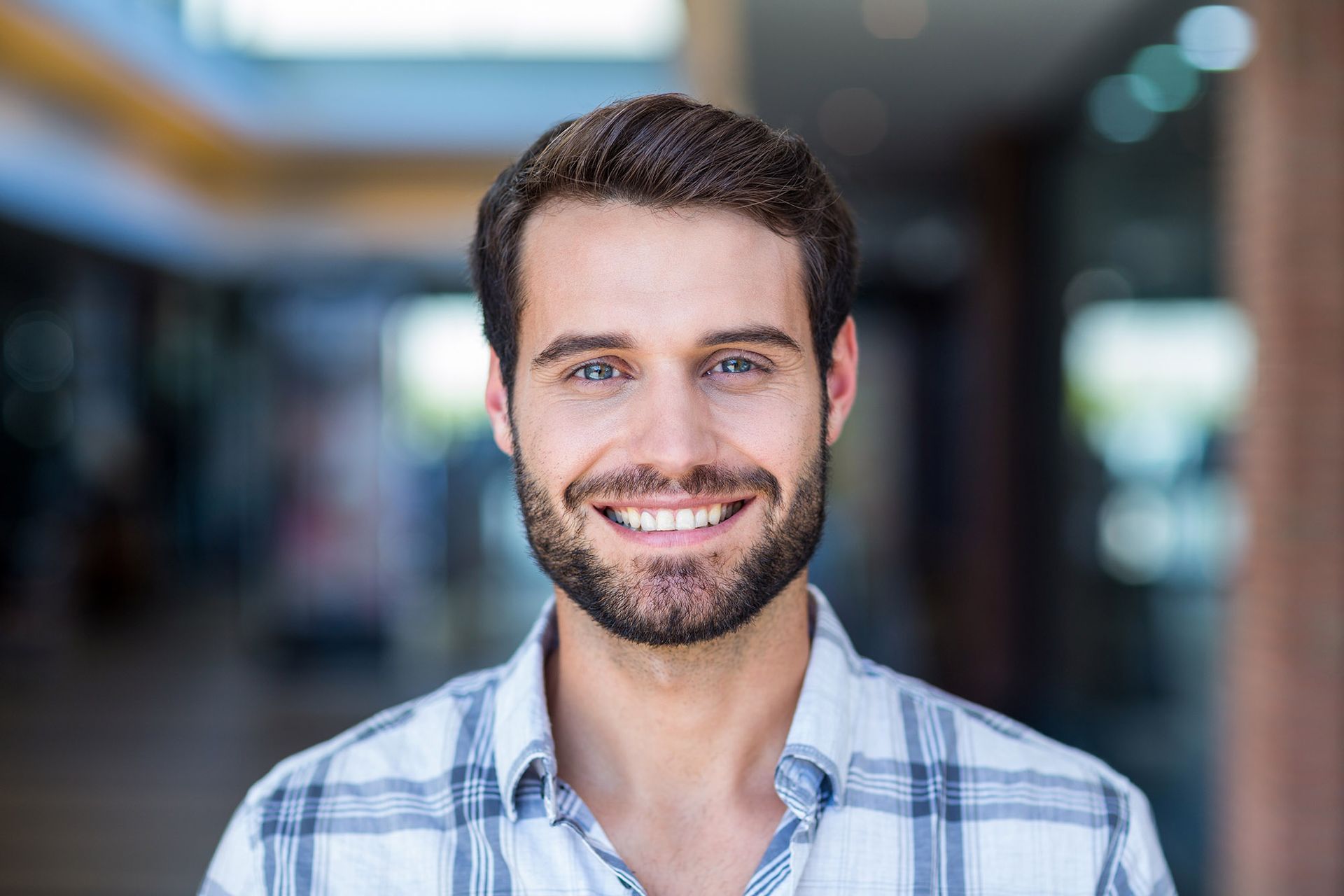 Man with blue eyes and a beard smiles at the camera in a building with a checkered shirt.