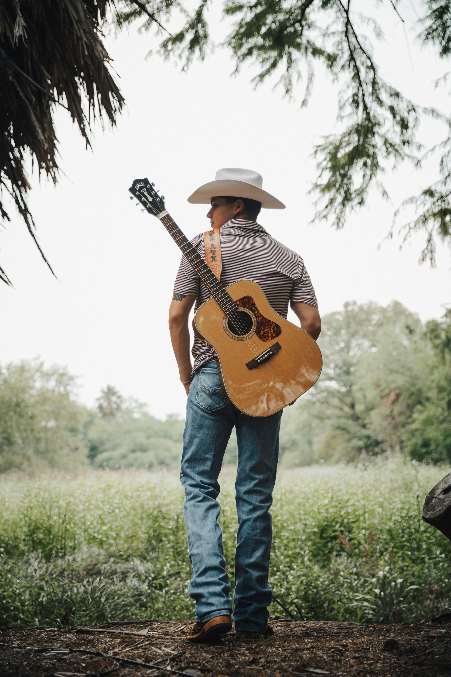 Man in cowboy hat and jeans, carrying guitar on his back, standing in a field.