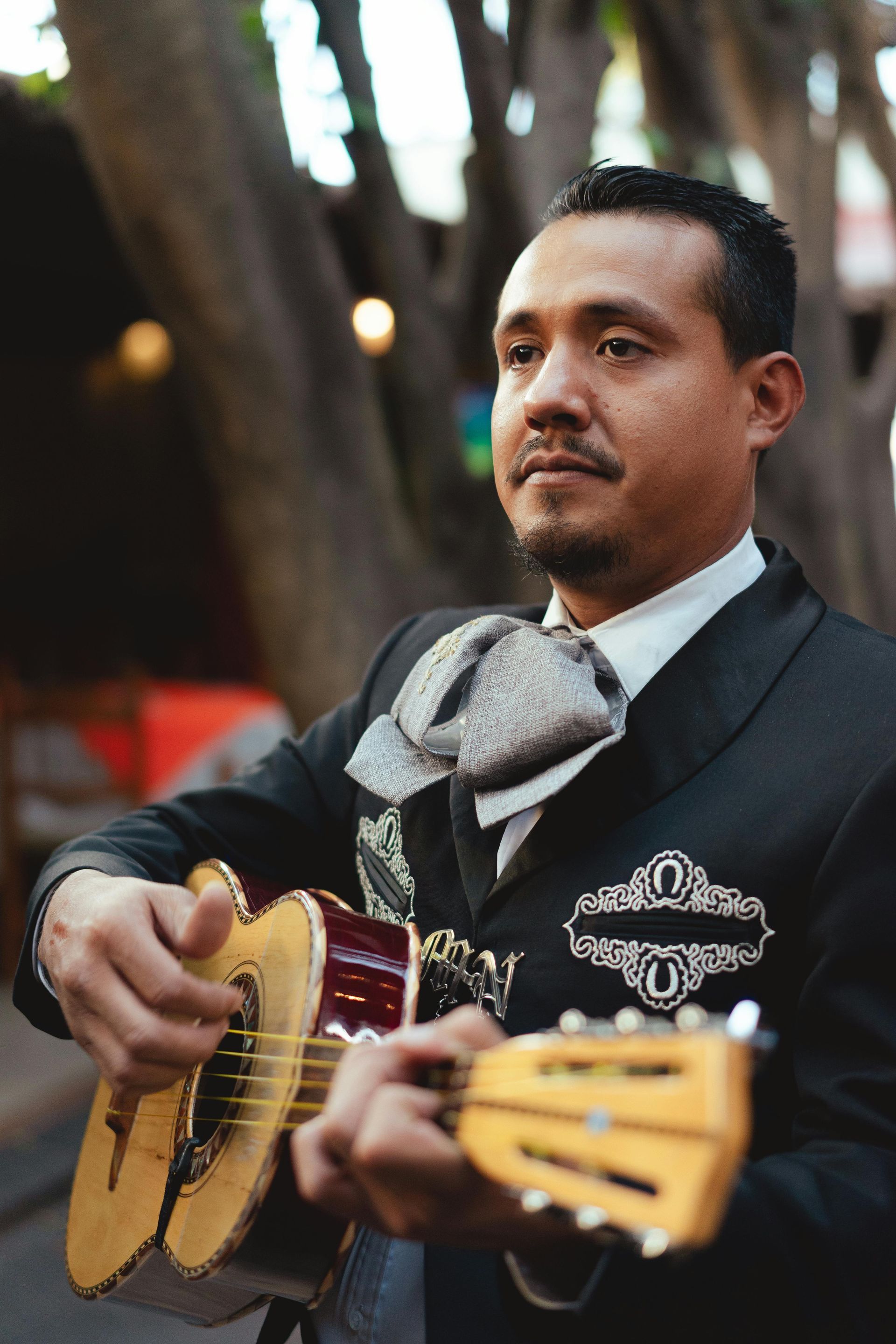 Man in mariachi attire playing a small guitar outdoors, gazing intently. He wears a black suit with ornate details, a large bow tie, and a serious expression.