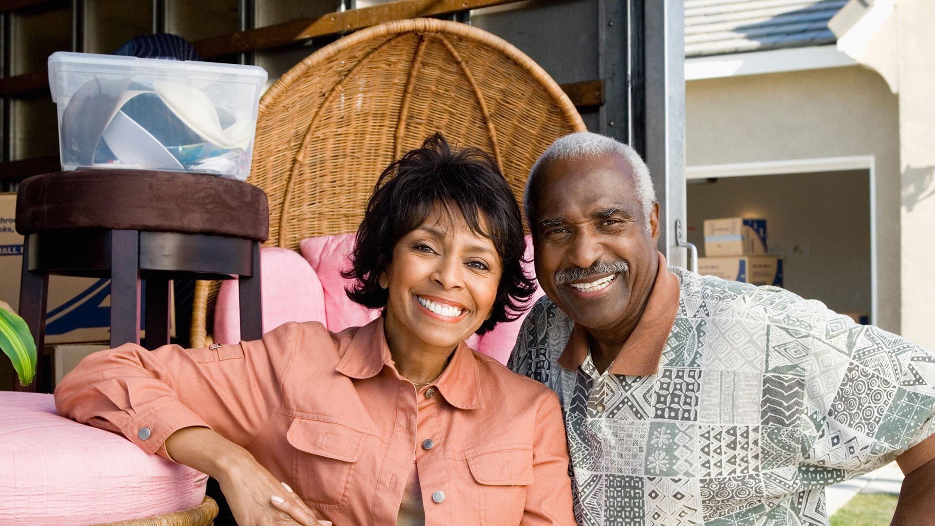 Smiling couple sitting on a porch with colorful cushions and a wicker chair behind them