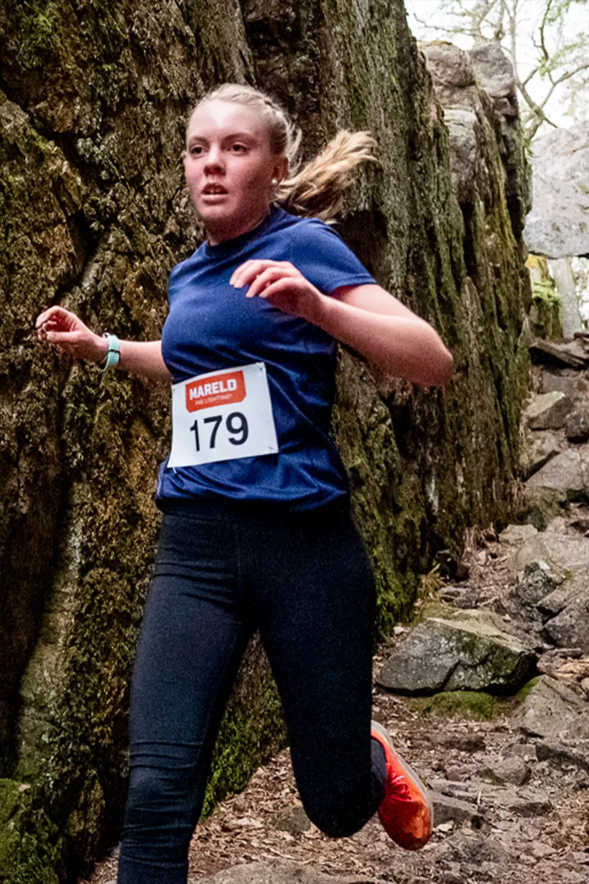 A woman in a blue shirt and black pants is running on a trail.