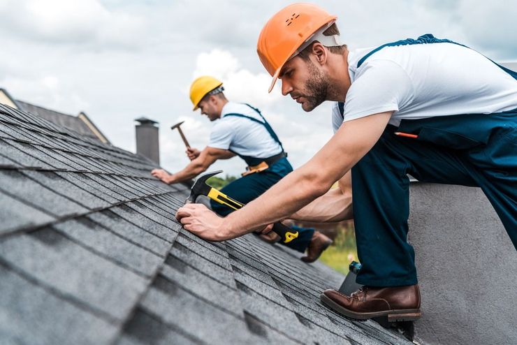 Roof workers installing shingles on a sloped roof, wearing hard hats and safety gear.