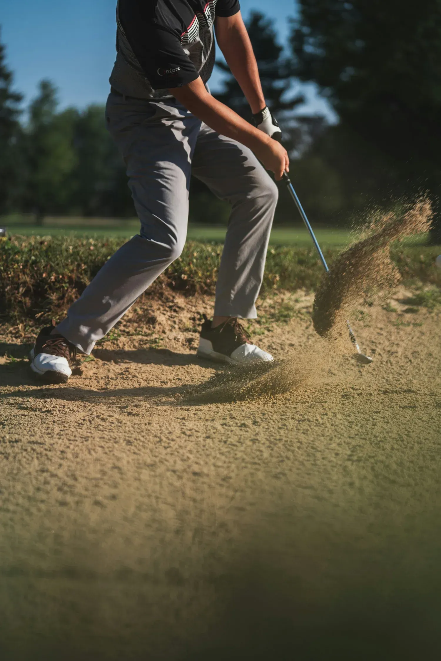 Ein Golfer schlägt aus einem sandigen Bunker ab, wobei Sand in die Luft spritzt.
