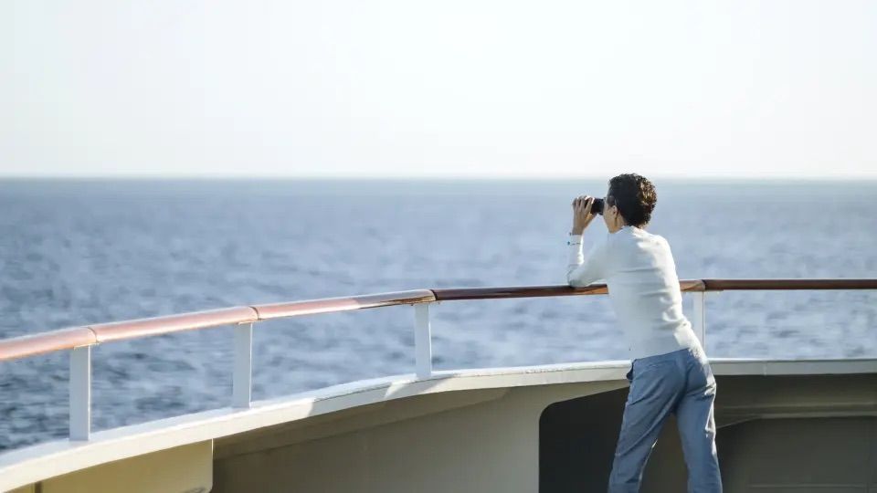 Eine Person steht an Deck eines Schiffes und blickt durch ein Fernglas auf das offene Meer.