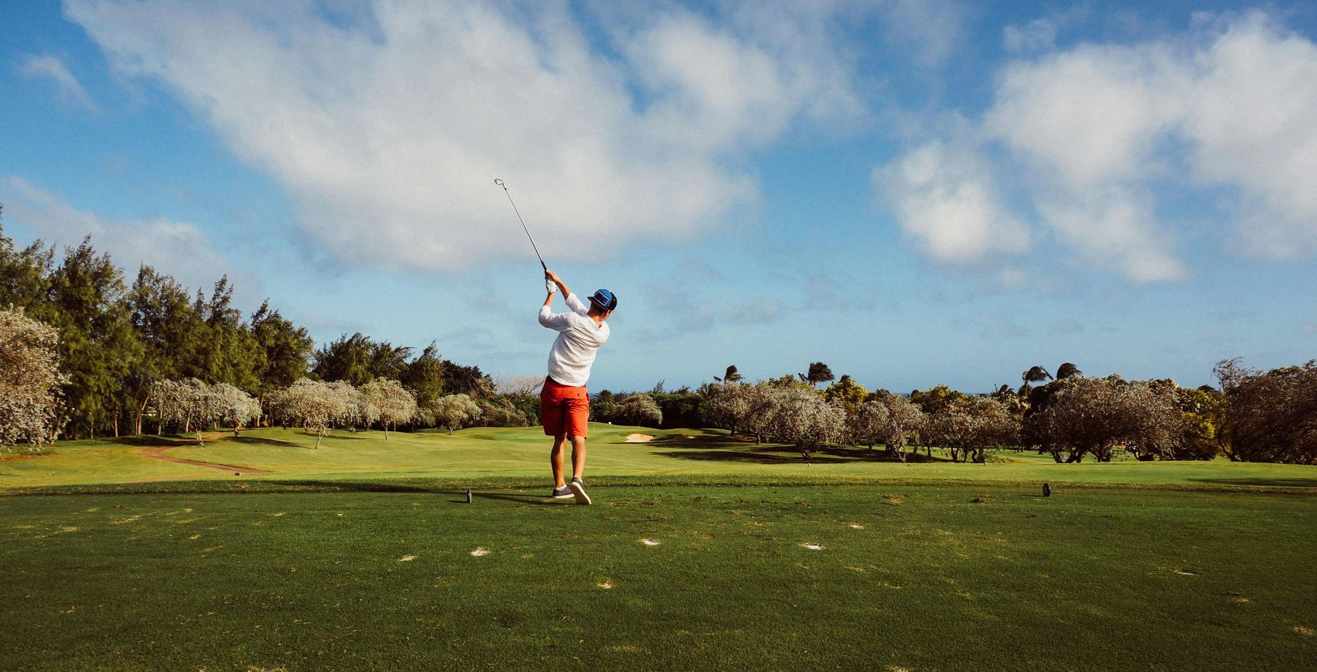 Ein Golfer in roten Shorts schwingt einen Schläger auf einem grünen Fairway unter blauem Himmel mit Wolken.