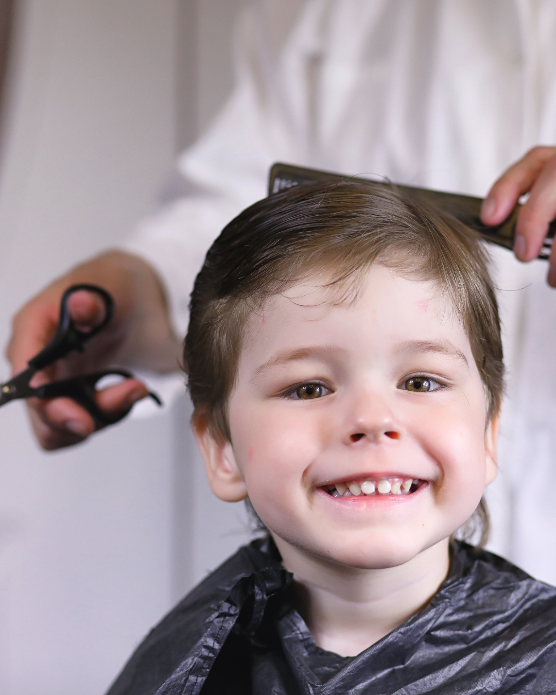 Un enfant sourit pendant qu'on lui coupe les cheveux. Une personne tient des ciseaux et un peigne.