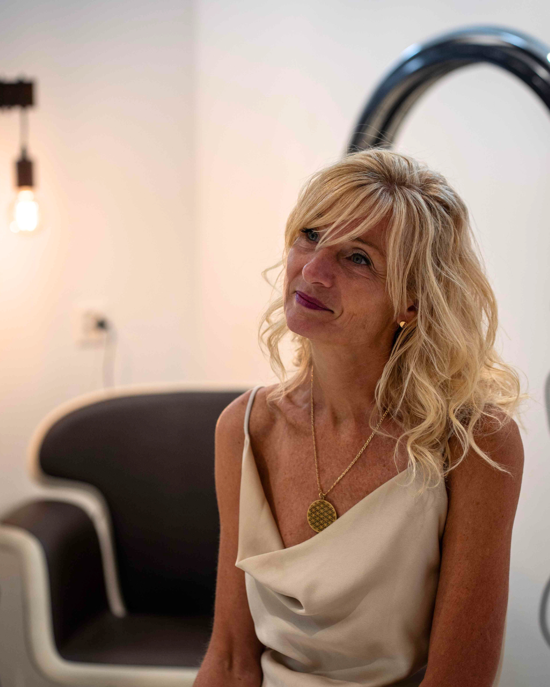 Femme vêtue d'un haut couleur crème, regardant vers le haut, portant un collier pendentif, assise près d'une chaise noire et blanche.