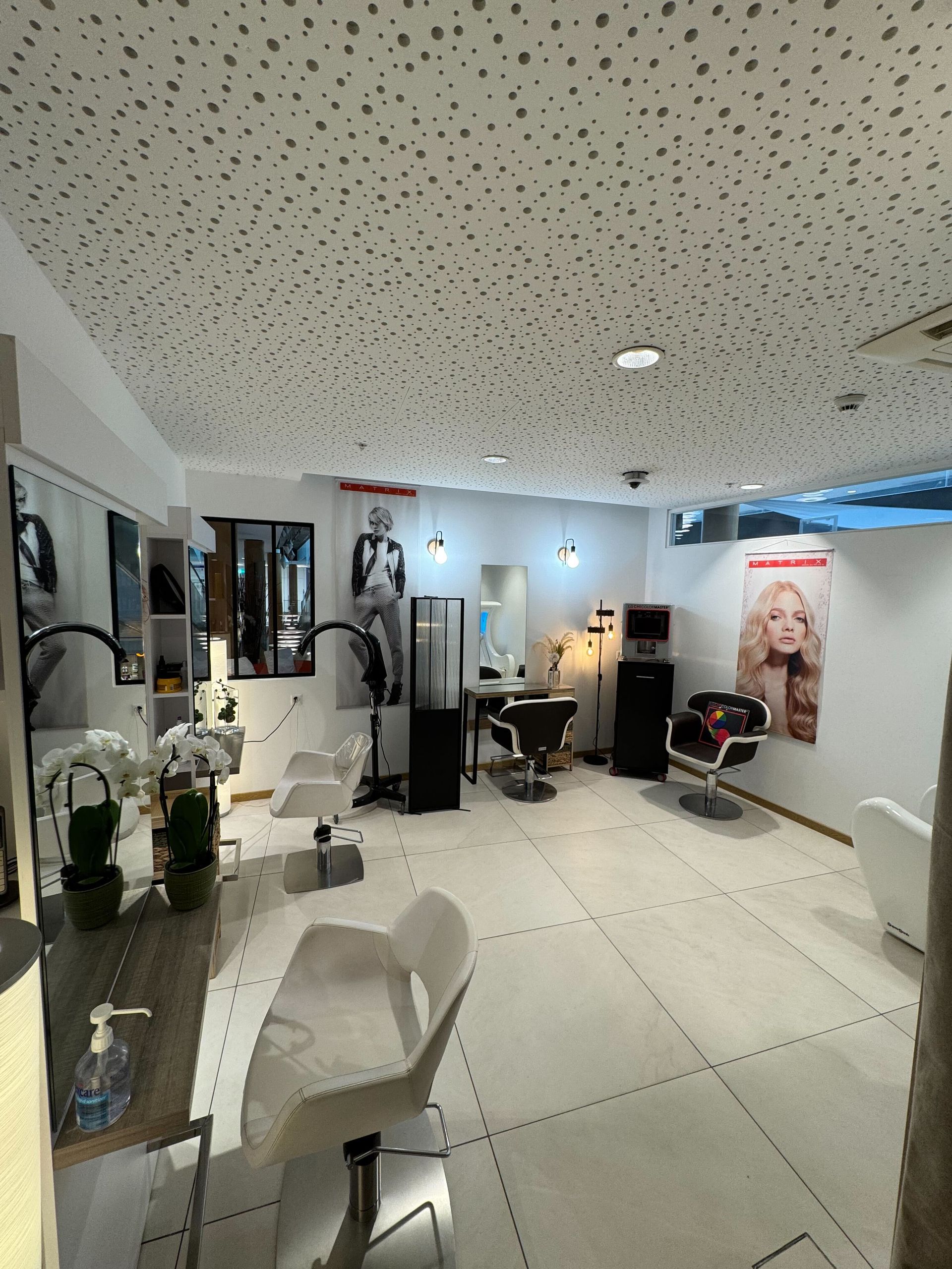 Intérieur d'un salon de coiffure avec des chaises blanches, des miroirs et un sol carrelé blanc.