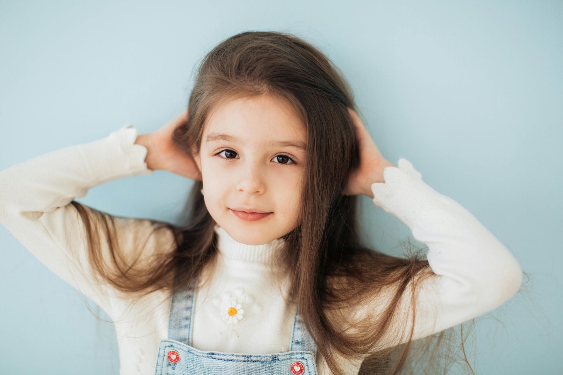 Jeune fille aux longs cheveux bruns, souriante, les mains dans les cheveux, vêtue d'un haut blanc et d'une salopette en jean, sur fond bleu.