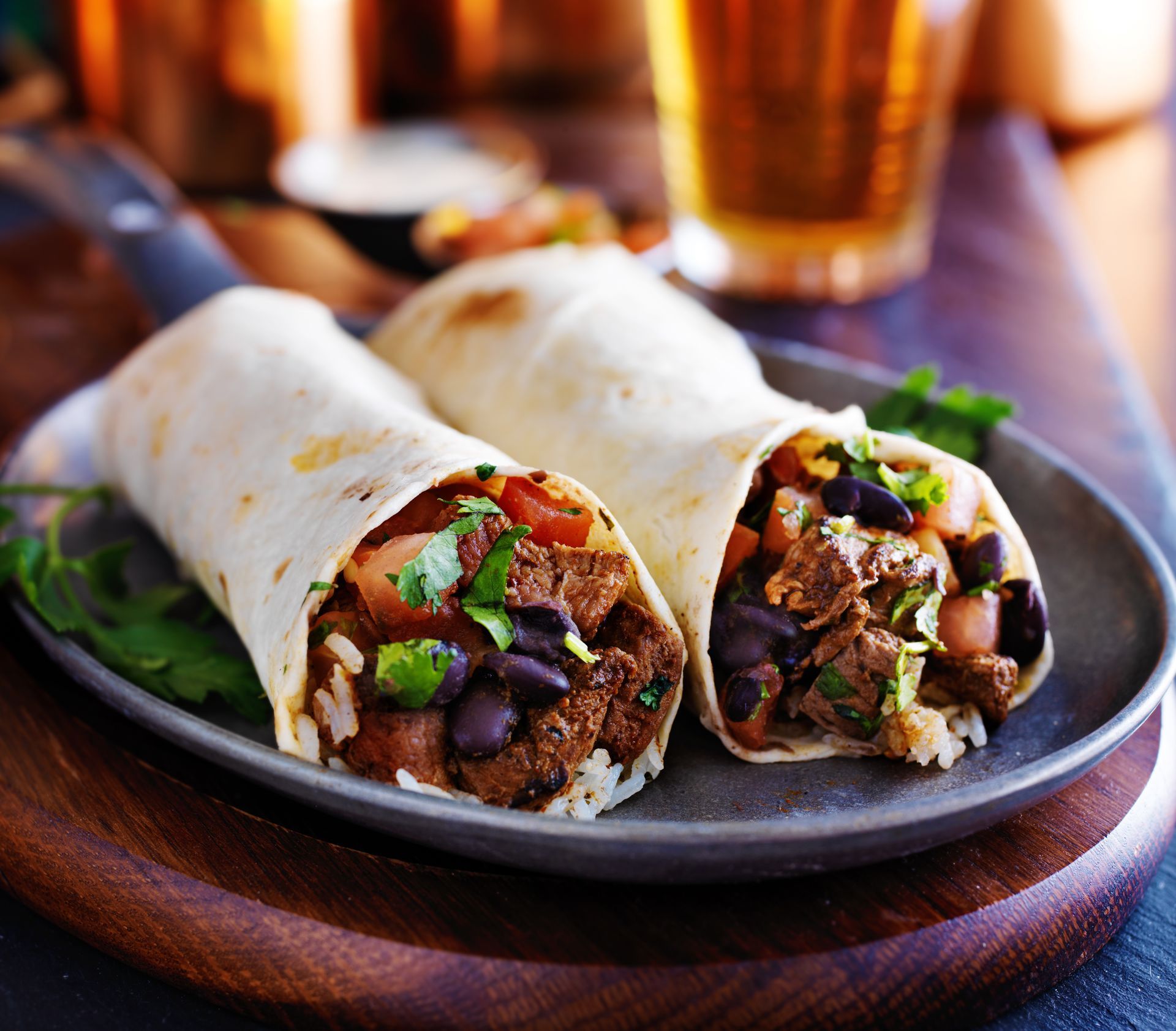 Two burritos are on a plate with a glass of beer in the background.