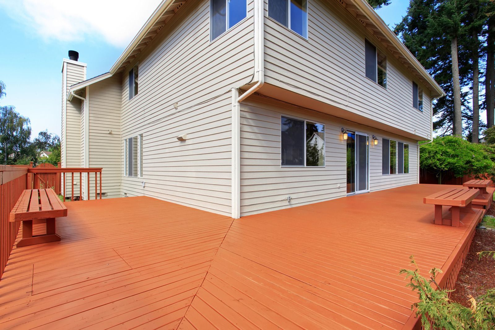 Two-story house with a large wooden deck painted orange-brown. Sunny day, with trees in background.